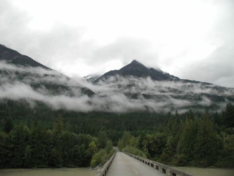 1990 Ford F-250 - A bridge crosses a muddy river leading towards a dense evergreen forest at the base of mist-shrouded mountains under a cloudy sky.