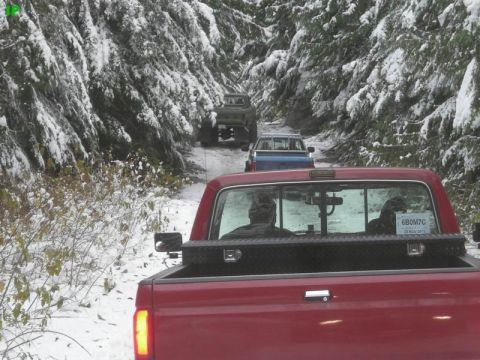 1990 Ford F-250 - A red Ford pickup truck is viewed from the rear, driving on a snow-covered road through a forest. Two other trucks are visible ahead on the…