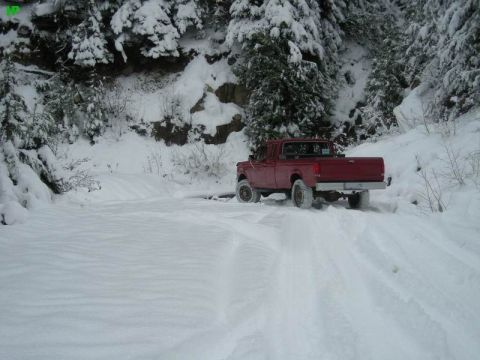 1990 Ford F-250 - A red Ford F-250 pickup truck drives on a snow-covered road surrounded by snow-laden trees. The truck is viewed from the rear quarter angle…