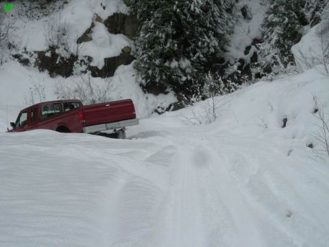 1990 Ford F-250 - A red Ford F-250 pickup truck is stuck in deep snow on a wooded hillside. The truck is angled into a snowdrift, with its rear end visible.