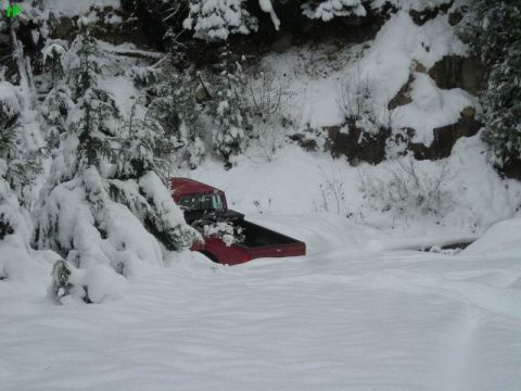 1990 Ford F-250 - A red Ford pickup truck is partially buried in deep snow on a wooded mountainside. The truck is angled away from the viewer, with its bed f…