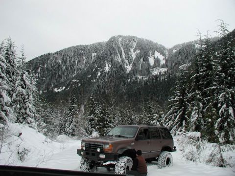 1990 Ford F-250 - A brown Ford Explorer SUV is parked in a snowy forest clearing with a person kneeling beside the rear tire. The vehicle has large off-road…