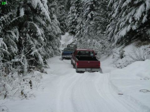 1990 Ford F-250 - A red Ford F-series pickup truck drives down a snow-covered road surrounded by snow-laden evergreen trees. Another blue pickup truck is vis…