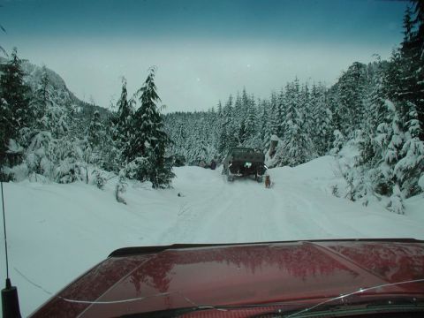 1990 Ford F-250 - The front of a red Ford F-250 is visible in the foreground, with a snow-covered forest and a military-style truck in the background.