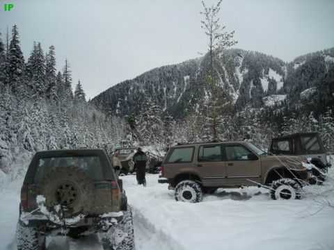 1990 Ford F-250 - Several off-road vehicles are parked in a snowy forest with snow-covered mountains in the background. The vehicle in the foreground is a br…