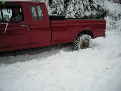 1990 Ford F-250 - A red Ford F-250 pickup truck is stuck in deep snow, with the driver visible behind the wheel. The truck's rear tire is partially submerged…