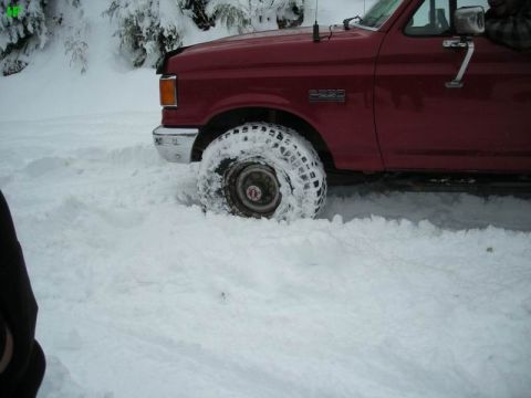 1990 Ford F-250 - A red Ford F-250 pickup truck is stuck in deep snow, with its front tire partially buried. The scene is set in a snowy, wooded area.