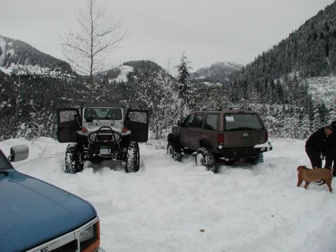 1990 Ford F-250 - Two off-road vehicles, a white Jeep and a brown SUV, are parked in deep snow with mountains and snow-covered trees in the background. A tan…