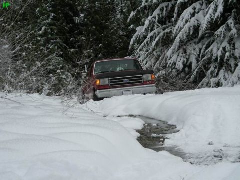 1990 Ford F-250 - A red Ford F-250 pickup truck is partially submerged in a snowy creek bed, surrounded by snow-covered trees.