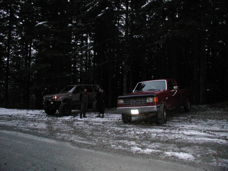 Two off-road vehicles, a red pickup and a dark SUV, parked in a snowy forest at dusk.