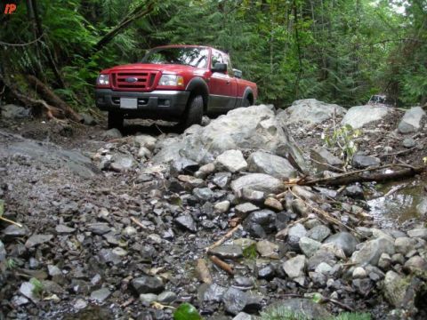 1990 Ford F-250 - A red Ford pickup truck navigates a rocky, off-road trail in a dense forest. The truck is angled towards the viewer, with its front end vis…