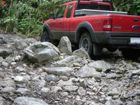 1990 Ford F-250 - A red Ford pickup truck navigates a rocky, off-road trail surrounded by dense foliage. The truck's rear is visible, showing its off-road ti…
