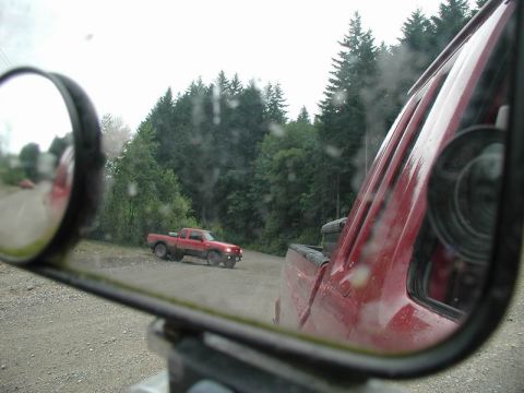 1990 Ford F-250 - A red pickup truck is seen in the side mirror of another red pickup truck, parked on a gravel road with a forest in the background.