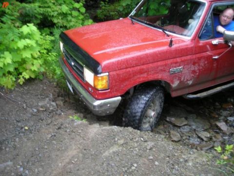 1990 Ford F-250 - A red Ford F-250 pickup truck is navigating a rough, rocky trail with its front passenger side tire stuck in a ditch. The truck is partiall…