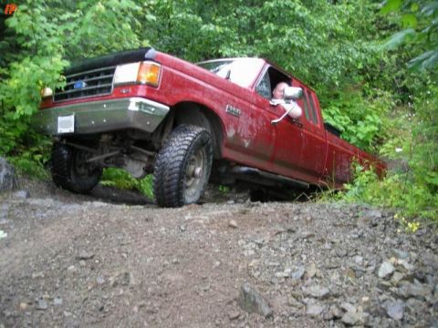 1990 Ford F-250 - A red Ford pickup truck is driving off-road on a dirt path surrounded by lush green foliage. The driver's side door is open and a person is…