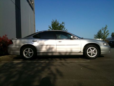 2002 Pontiac Grand Prix - The side profile of a silver 2002 Pontiac Grand Prix is shown parked in a lot next to a building. The car has tinted windows and five-spoke…