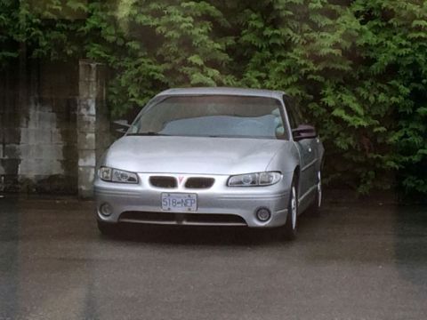 2002 Pontiac Grand Prix - The front view of a silver 2002 Pontiac Grand Prix is parked on a wet asphalt surface in front of a dense green hedge. The license plate re…