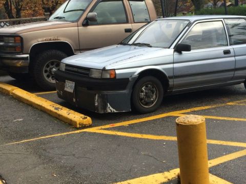 2002 Pontiac Grand Prix - A light blue hatchback is parked next to a brown pickup truck in a parking lot. The hatchback has a custom bumper and visible damage on its…