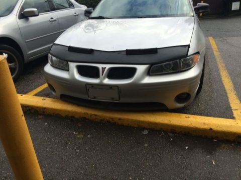2002 Pontiac Grand Prix - The front of a silver Pontiac Grand Prix is shown parked in a parking spot with a yellow curb. The car has a black car bra installed on the…