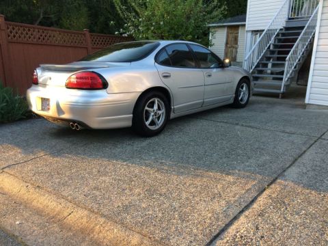 2002 Pontiac Grand Prix - The rear driver's side of a silver 2002 Pontiac Grand Prix is parked on a concrete driveway next to a house with an outdoor staircase.