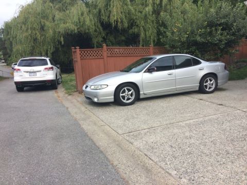 2002 Pontiac Grand Prix - A silver Pontiac Grand Prix is parked on a concrete driveway next to a wooden fence. A white SUV is parked on the side of the road behind i…