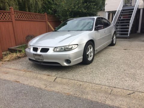 2002 Pontiac Grand Prix - A silver 2002 Pontiac Grand Prix is parked on a concrete driveway next to a wooden fence and a set of outdoor stairs. The front driver's si…