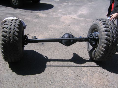 1978 Ford Bronco - A freshly black-painted solid front axle assembly standing on its own in a paved driveway, wearing mud-terrain tires and showing the offset…