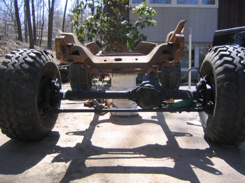 1978 Ford Bronco - Head-on view of a bare, surface-rusted truck frame on a concrete driveway, showing the front Dana-style solid axle with disc brakes, tie ro…