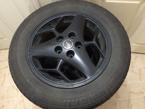 2002 Pontiac Grand Prix - A black Pontiac alloy wheel with a tire is shown in a close-up, angled shot. The Pontiac logo is visible in the center of the wheel.