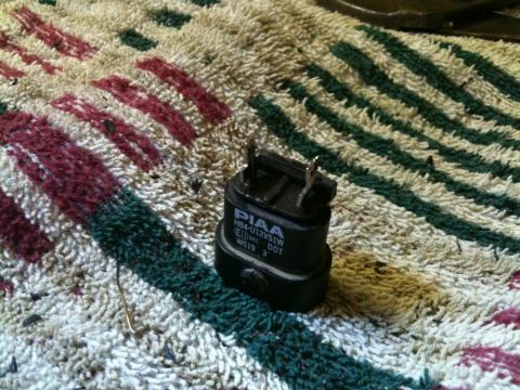 2002 Pontiac Grand Prix - A close-up shot of a black PIAA HB4U12V51W automotive light bulb base with visible text and markings, resting on a patterned rug.