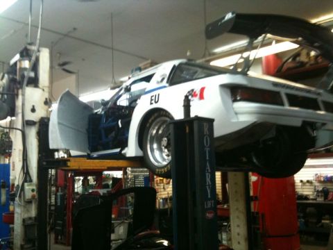 2002 Pontiac Grand Prix - A white race car with a large rear wing is elevated on a car lift in a repair shop. The driver's side door is open, revealing a blue roll c…