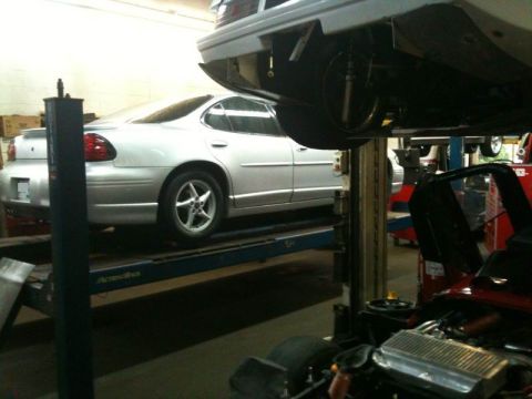 2002 Pontiac Grand Prix - A silver sedan is raised on a car lift in a garage. Another vehicle, partially visible in the foreground, appears to be a red sports car wi…
