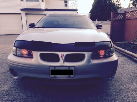 2002 Pontiac Grand Prix - The front view of a silver Pontiac Grand Prix with a black car bra is parked on a concrete driveway in front of a house. The headlights are…