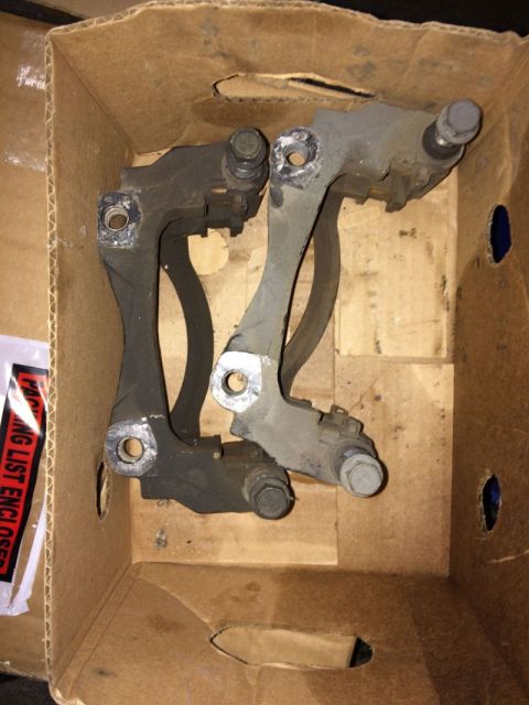 2002 Pontiac Grand Prix - Two used brake caliper brackets are shown resting inside a cardboard box. One bracket is black and the other is gray.