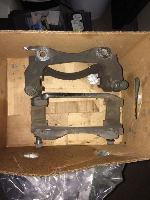 2002 Pontiac Grand Prix - Two black brake caliper brackets are shown inside a cardboard box. One bracket is stacked on top of the other, with bolts visible on both.