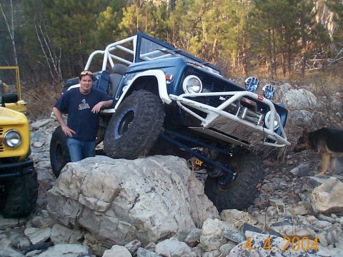 1977 Ford Bronco - Two modified off-road vehicles navigate a rocky trail with a man and a dog present.