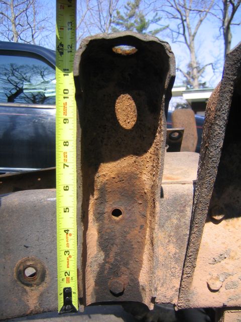 1978 Ford Bronco - A heavily rusted steel frame bracket held up against a yellow Stanley tape measure reading past 12 inches, photographed outdoors with an ol…