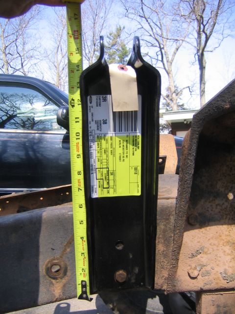1978 Ford Bronco - A black steel bracket with a Ford parts shipping label and barcode is held against a rusty vehicle frame, with a yellow tape measure alongs…