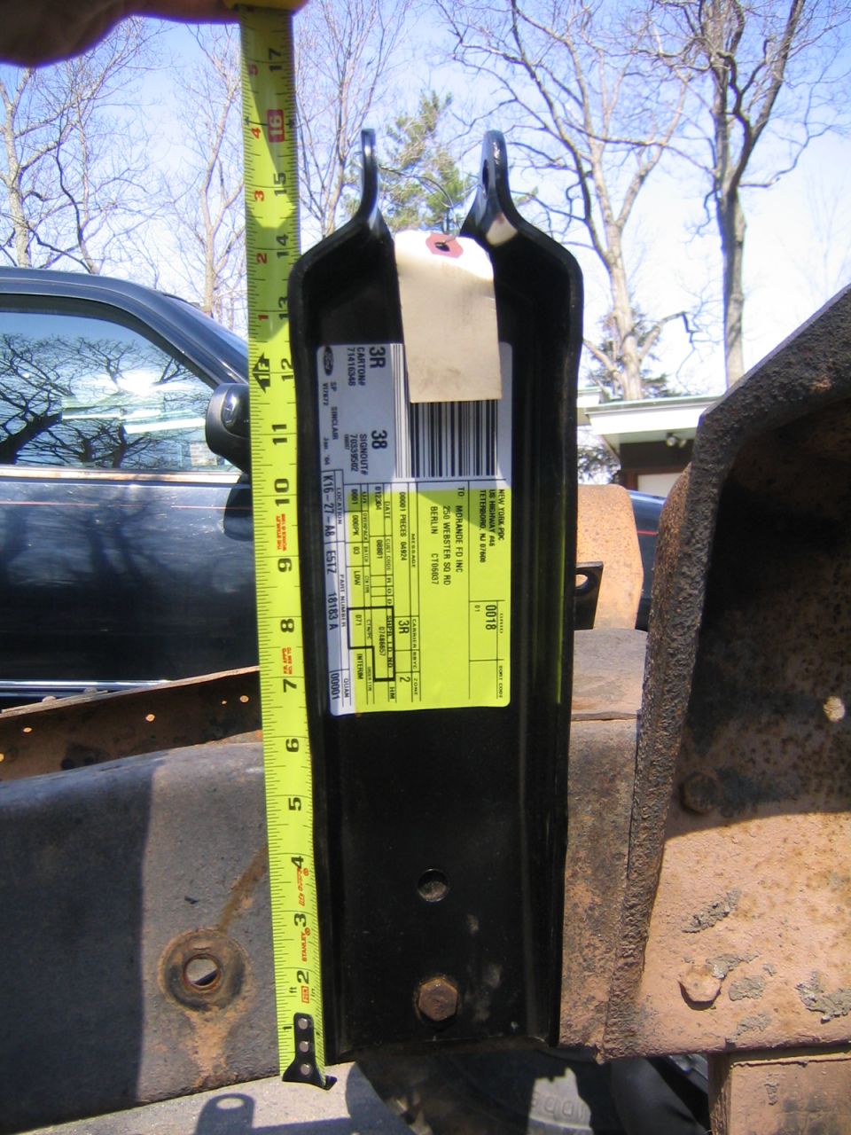 Black steel Ford part bracket with shipping label held against a rusty frame, tape measure for scale