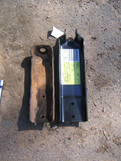 1978 Ford Bronco - Side-by-side comparison of an old rust-pitted stamped-steel shock mount bracket and its new gloss-black replacement, laid on gravel ground.…