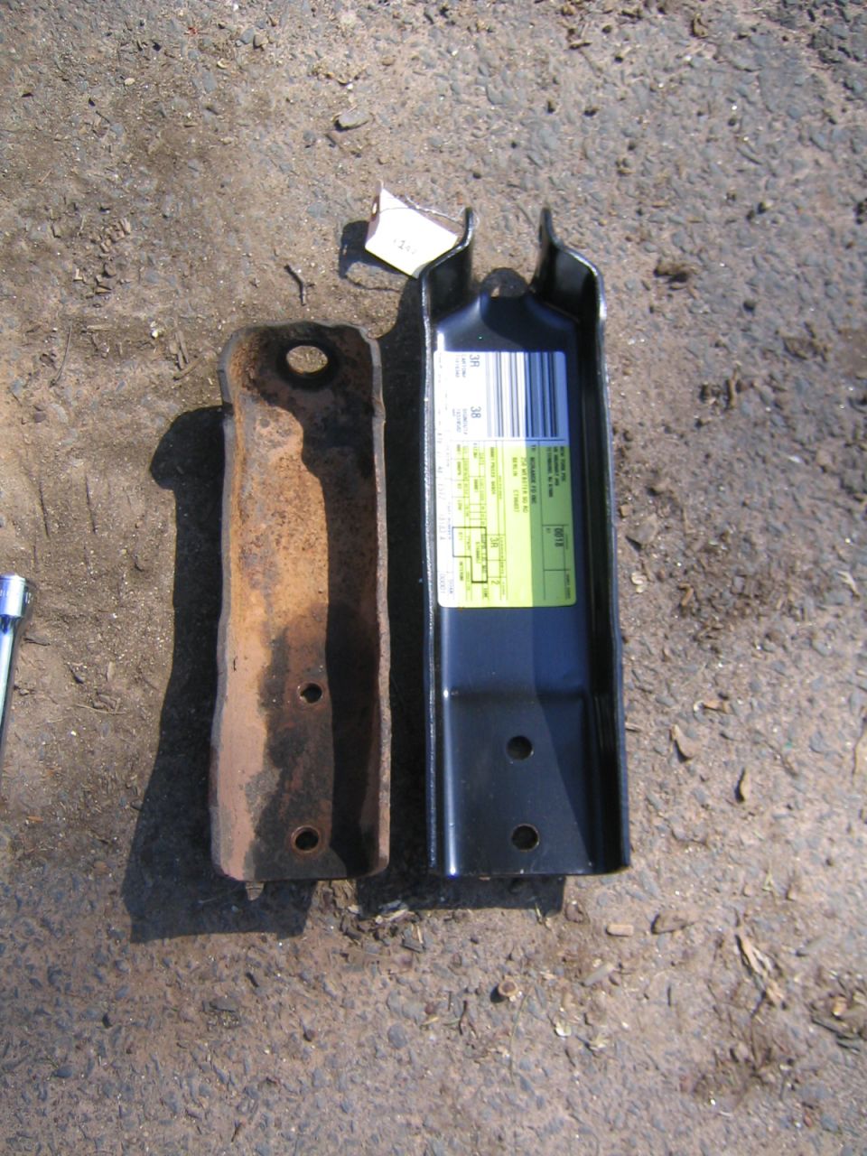 Rusty old shock mount bracket next to new black replacement bracket with parts label on gravel