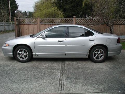 2002 Pontiac Grand Prix - The side view of a silver 2002 Pontiac Grand Prix is parked on a concrete driveway in front of a wooden lattice fence.