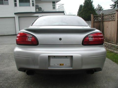 2002 Pontiac Grand Prix - The rear view of a silver Pontiac Grand Prix is shown parked on a paved driveway in front of a house. The car has a spoiler and dual exhaus…