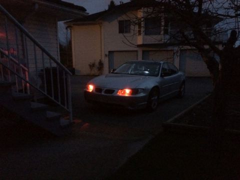 2002 Pontiac Grand Prix - A silver sedan with its headlights and parking lights illuminated sits in a driveway at dusk. The vehicle is parked in front of a residenti…
