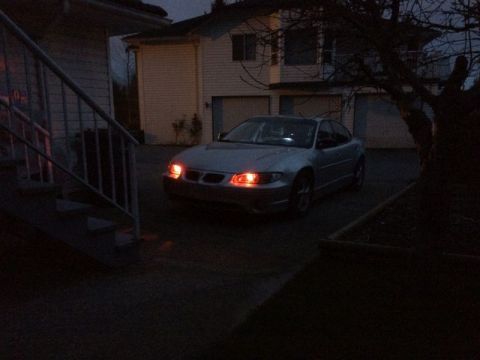 2002 Pontiac Grand Prix - A silver Pontiac Grand Prix sedan is parked in a driveway at dusk with its headlights and parking lights illuminated. The car is viewed fro…