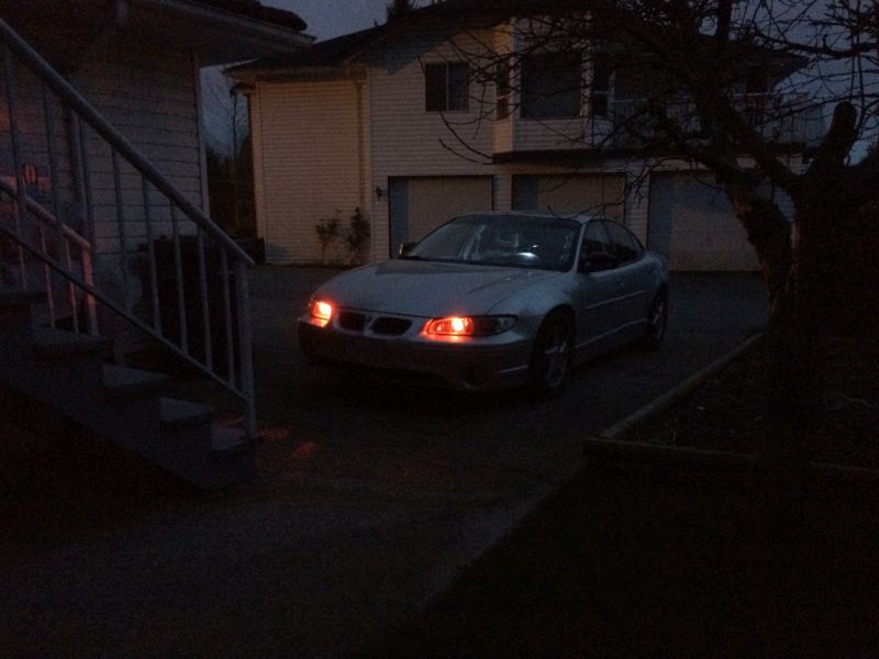 Silver 2002 Pontiac Grand Prix sedan with headlights on, parked in driveway at dusk.