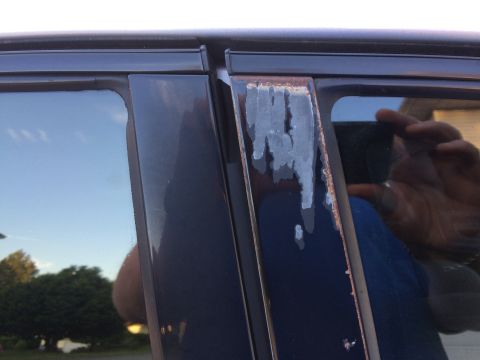 2002 Pontiac Grand Prix - The image shows a close-up of the B-pillar of a dark blue car, with visible rust and peeling paint on the trim. The window reflects the sky…