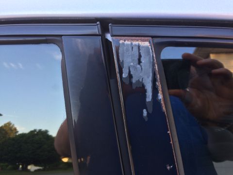 2002 Pontiac Grand Prix - Close-up of the driver's side B-pillar on a dark blue vehicle, showing significant rust and peeling paint along the edge of the window trim…