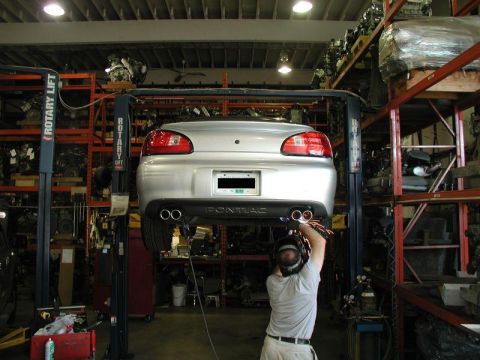 2002 Pontiac Grand Prix - A silver Pontiac Grand Prix is lifted on a car lift in a repair shop, with a mechanic working on the exhaust system. The car is viewed from…