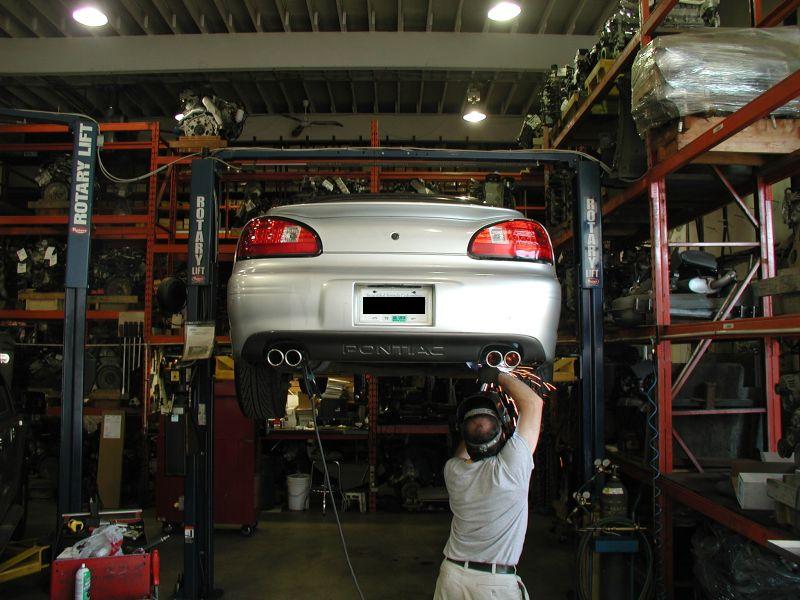 Silver Pontiac Grand Prix on a car lift, mechanic working on rear exhaust system.